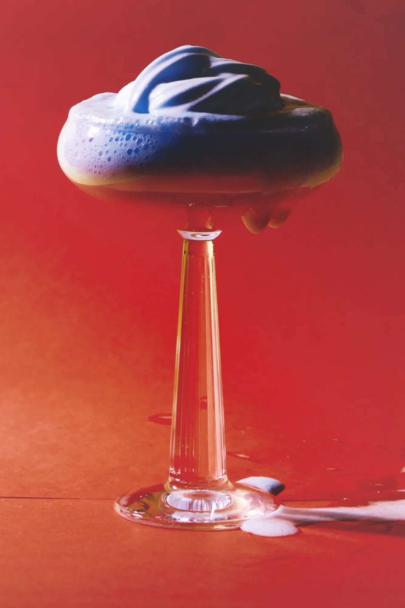 A coupe glass piled with blue-violet foam against a red wall, with foam spilled on the table