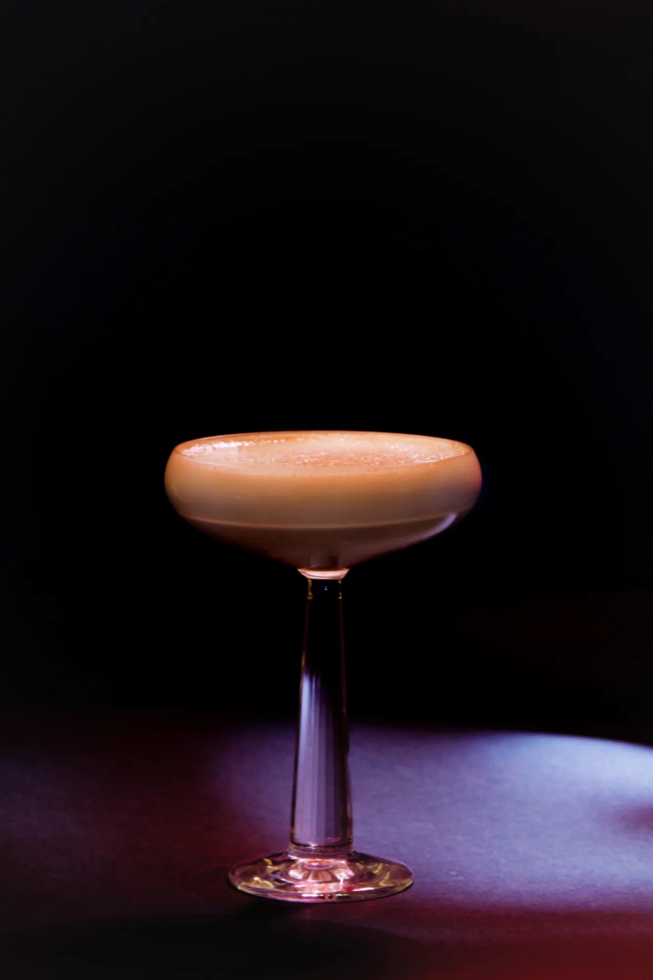 A pale foamy cocktail in a coupe glass, lit against a black background