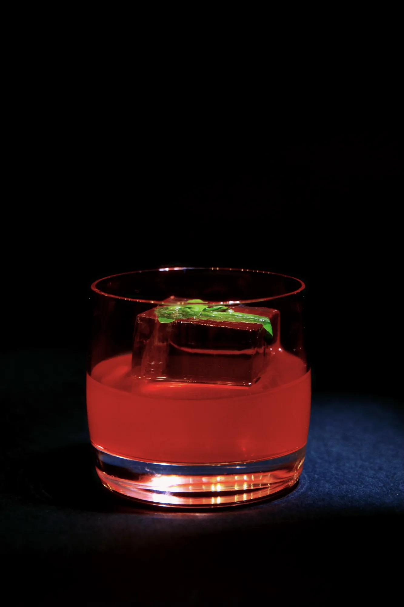 A red cocktail in a rocks glass with a large ice cube and fresh mint