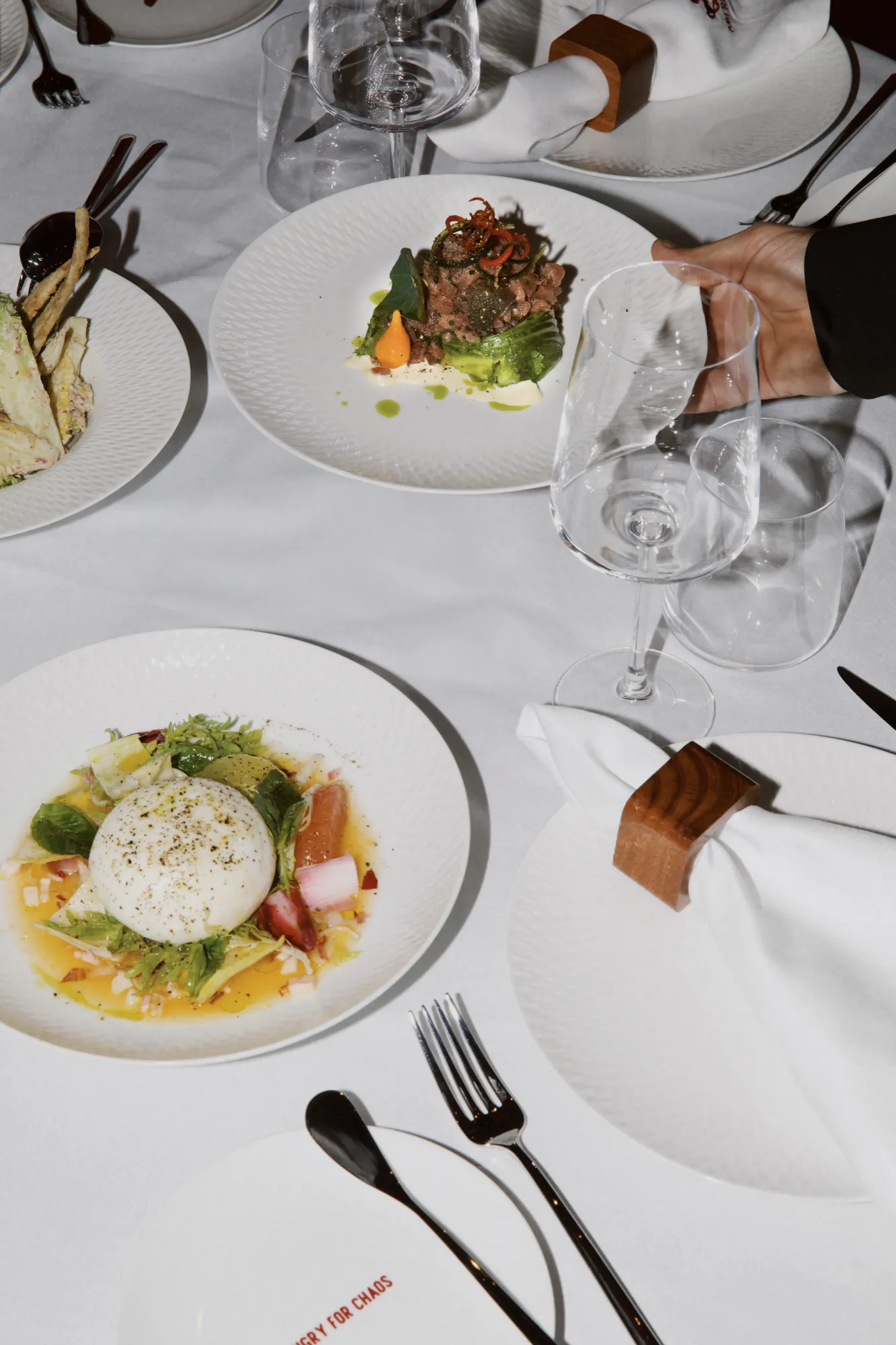 An overhead view of a table at Abi Rouge, with burrata, tartare, and a hand setting down a wine glass