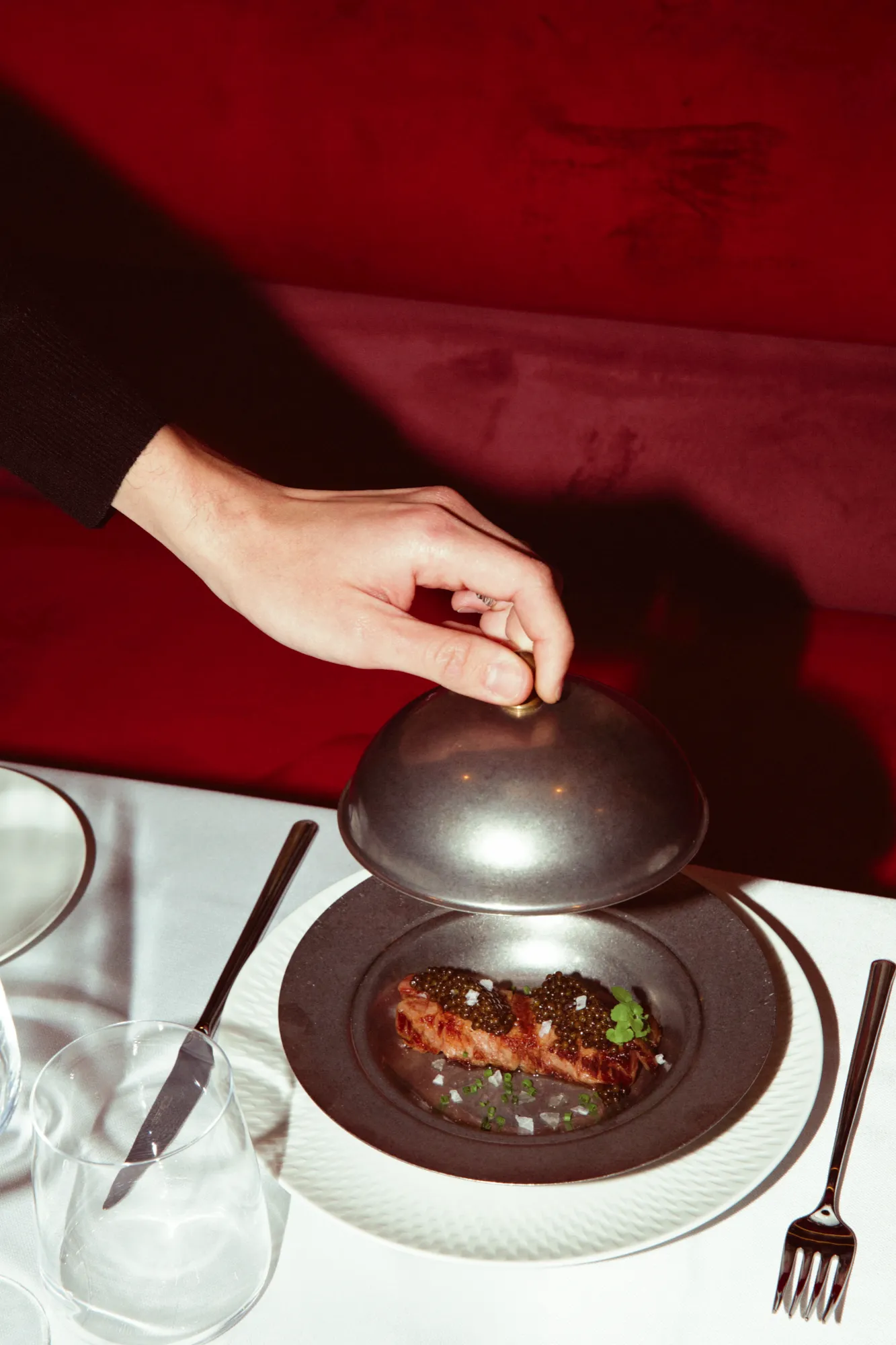 A hand lifting a silver cloche from a dish topped with caviar, in front of a red banquette
