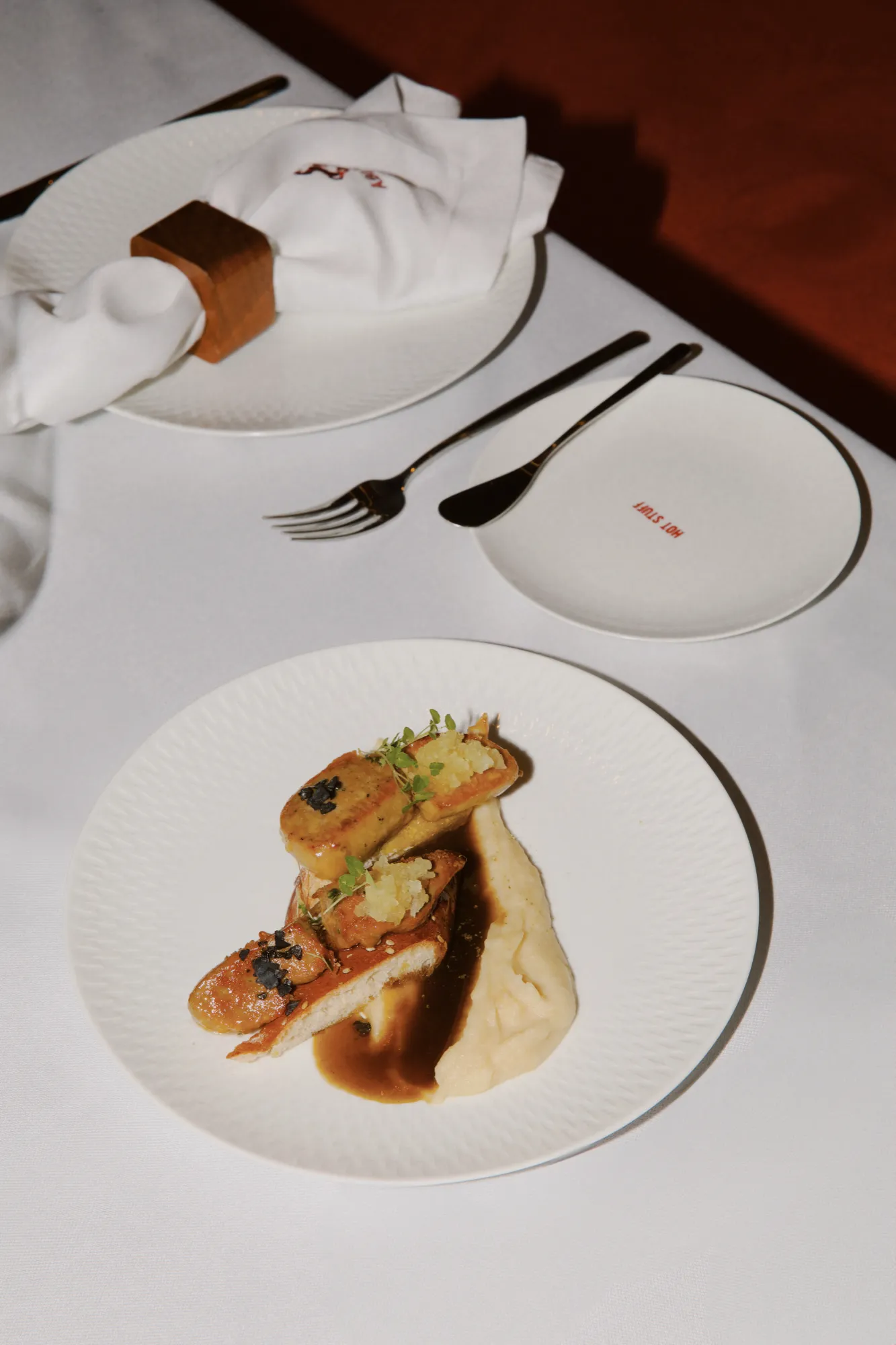 Glazed poultry with puree and herbs on a white plate, with cutlery and a folded napkin behind it