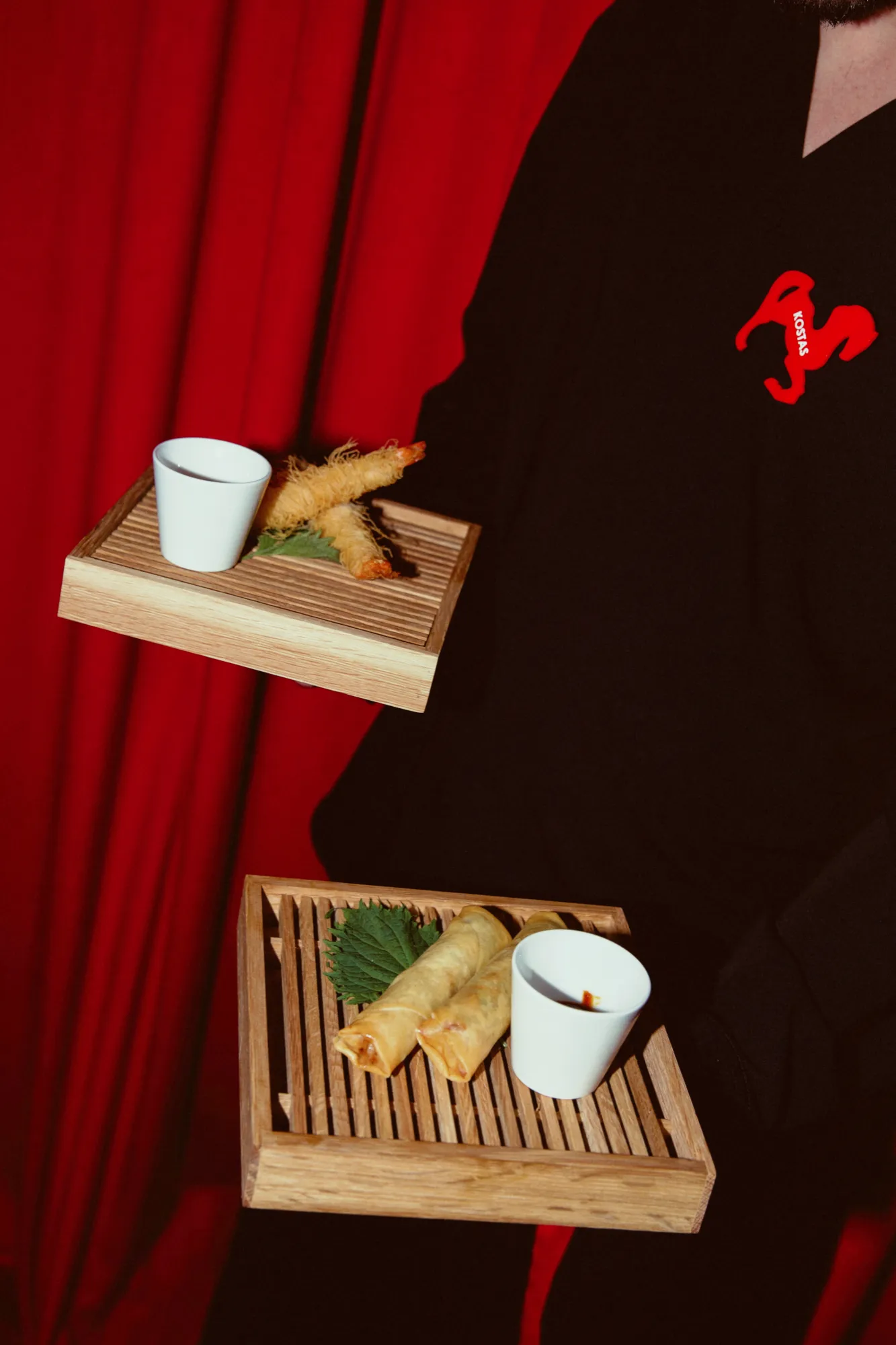 A server in black, with the red horse on the chest, carrying two wooden trays of spring rolls