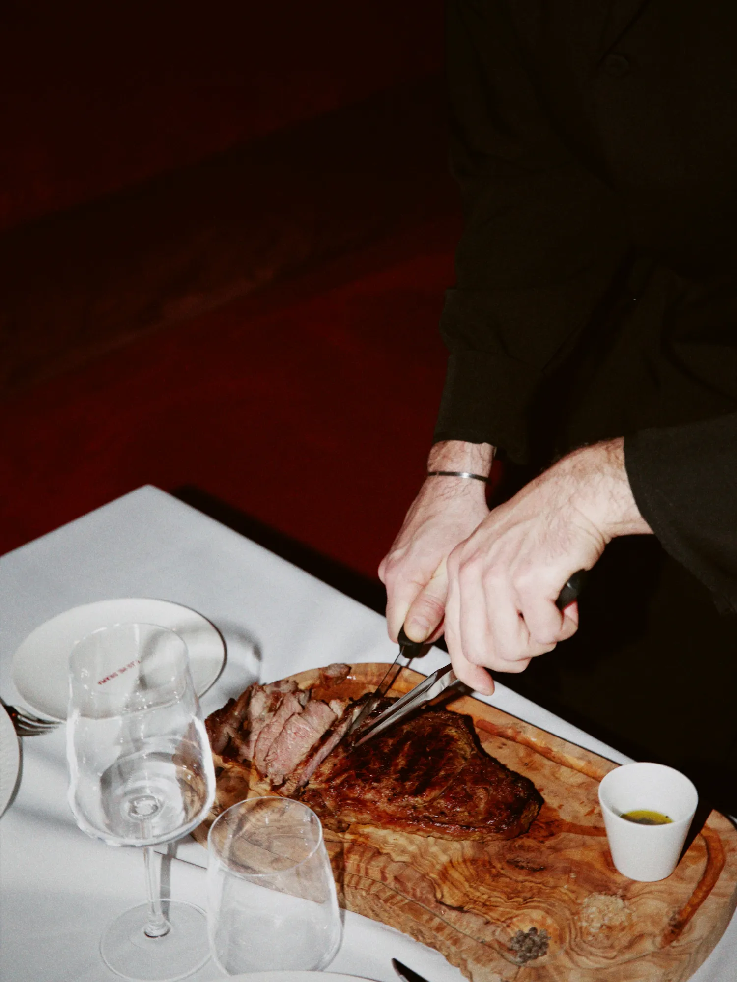 Hands carving a steak on a wooden board at a table set with wine glasses