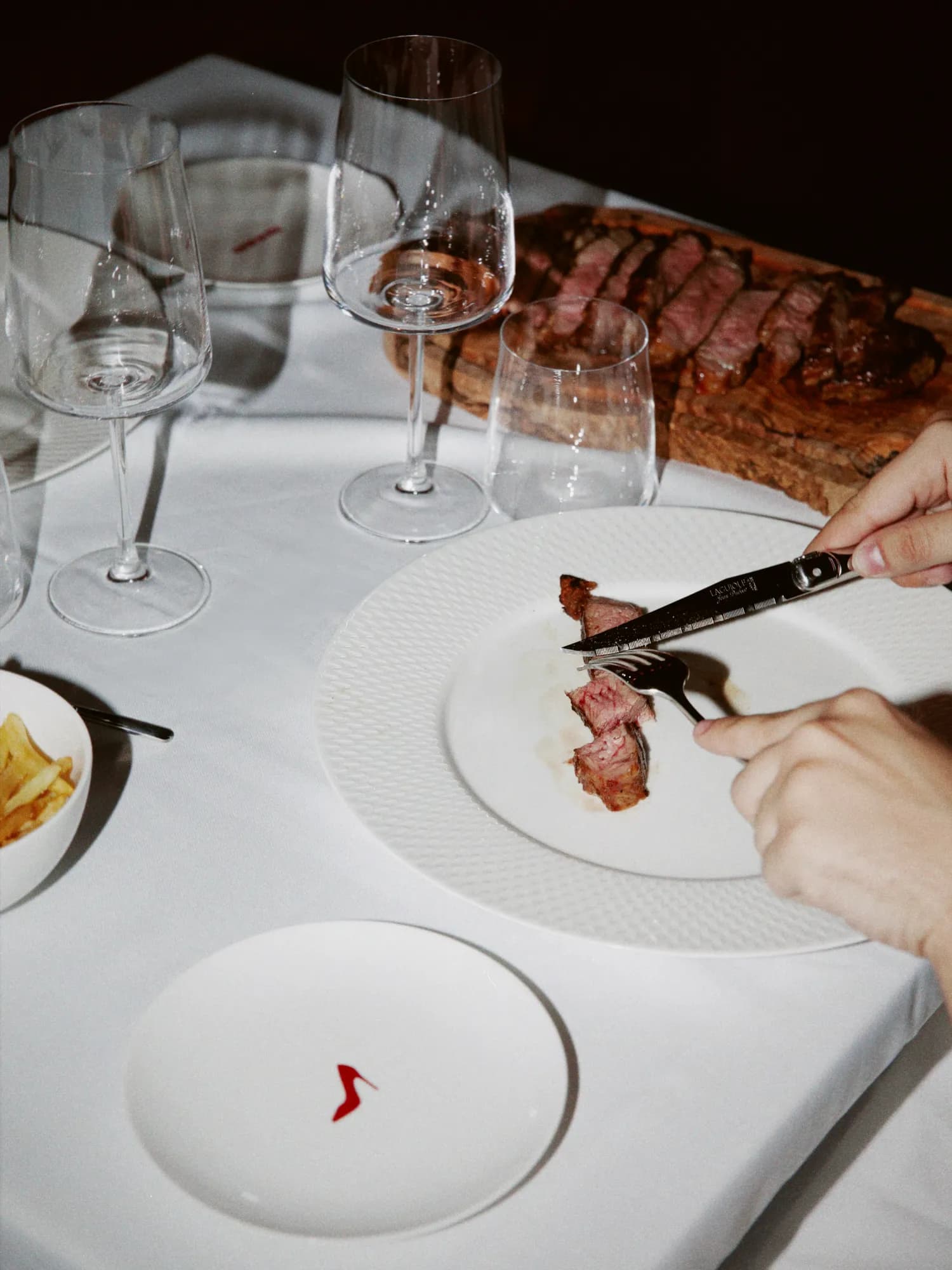 Sliced steak being served onto a white plate, with the carving board and wine glasses behind it