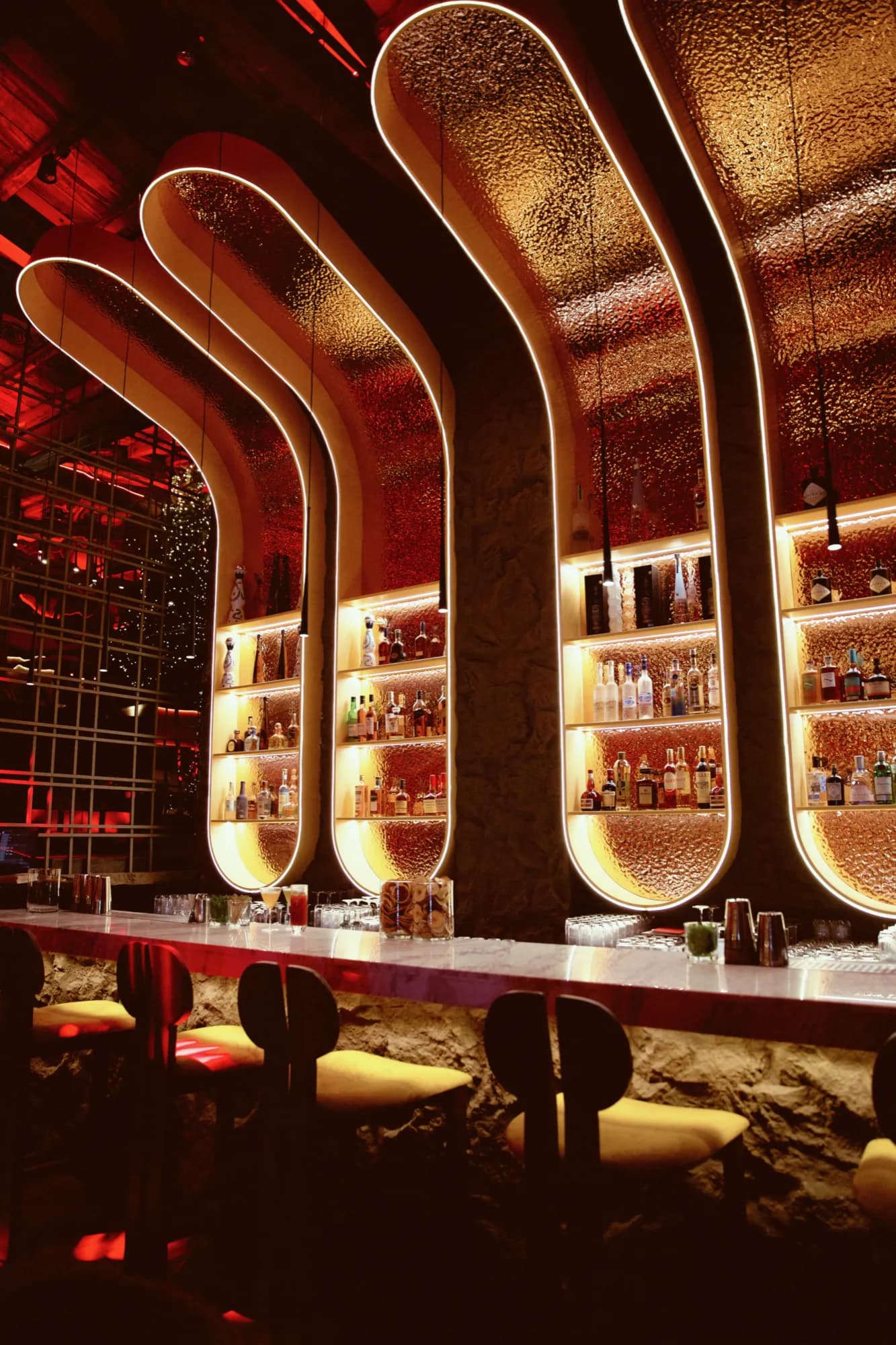 The bar, with glowing arched shelves of bottles and yellow stools along a marble counter