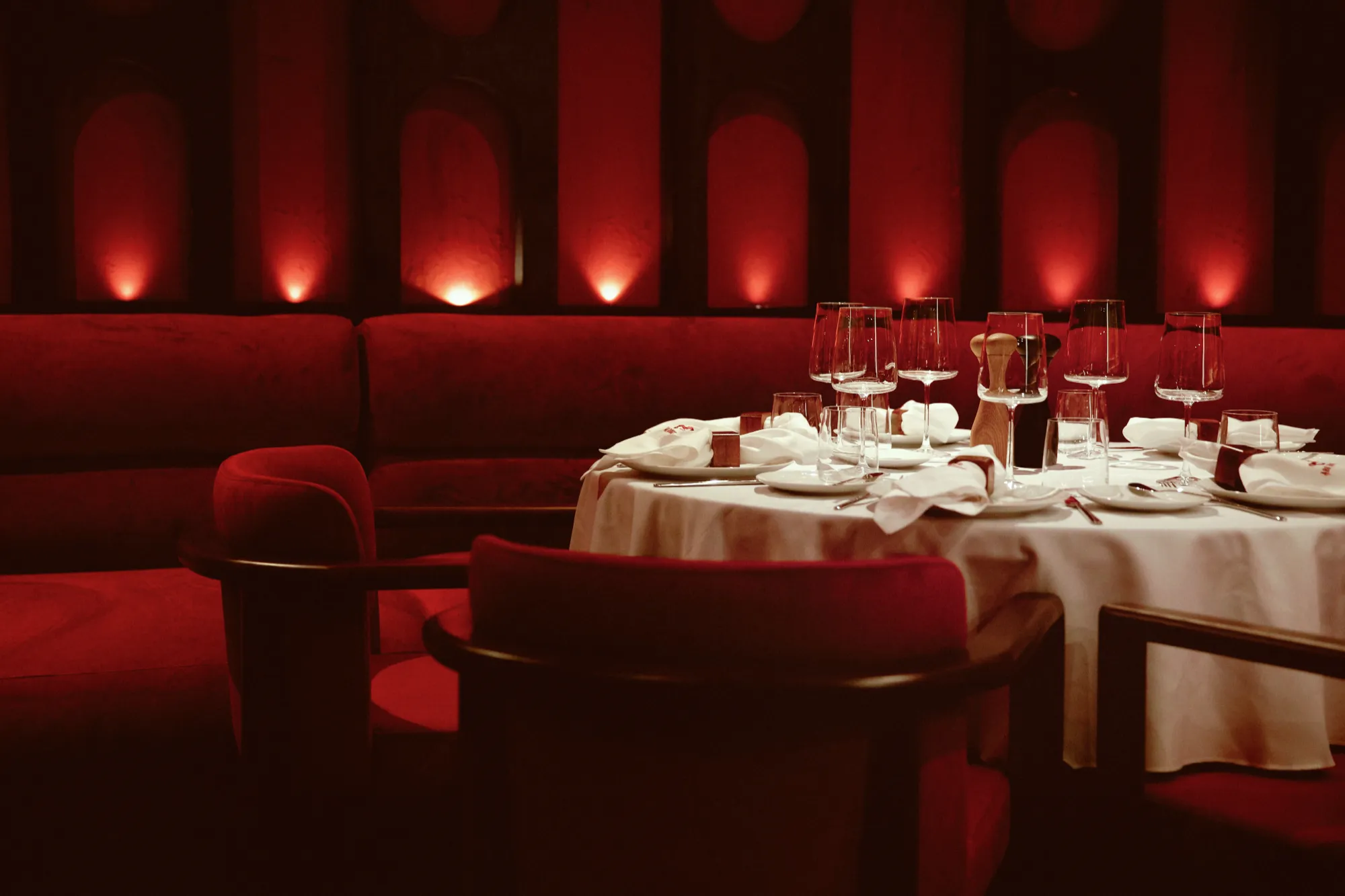 A round table set for dinner in a red banquette, with wine glasses against the lit wall